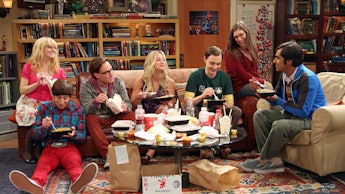 The cast of The Big Bang Theory sitting in the living room eating takeout in a scene of an episode