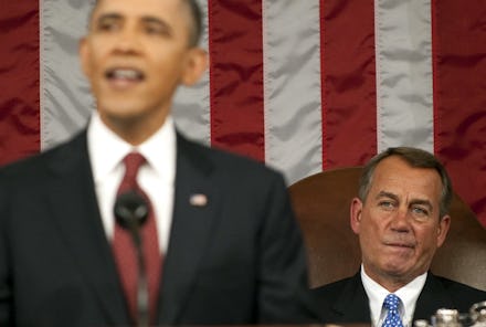 Barack Obama mid-sentence while giving a speech, with the American flag and another man in the back.