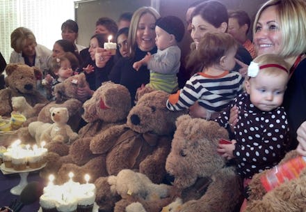 A group of women holding their children in their laps with teddy bears, and cupcakes with candles in...