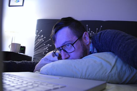 A man under 30 in glasses has his head on a pillow while looking at his laptop