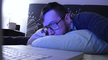 A man under 30 in glasses has his head on a pillow while looking at his laptop