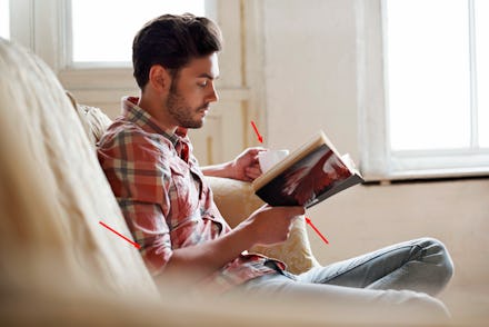 A man with brown hair sitting on a couch, drinking from a cup, and reading a book, and that makes hi...