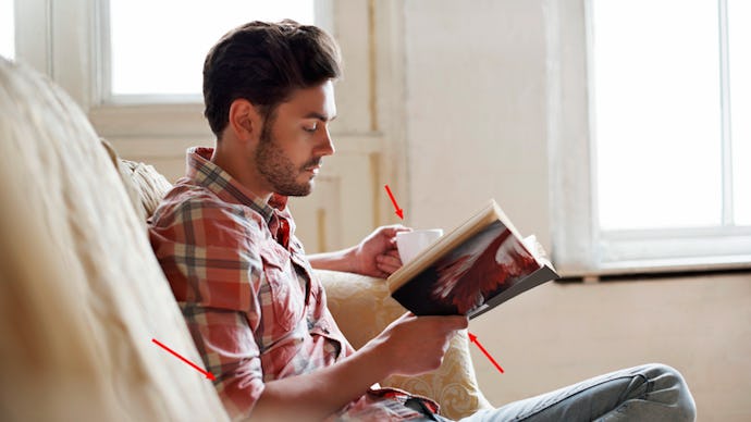 A man with brown hair sitting on a couch, drinking from a cup, and reading a book, and that makes hi...