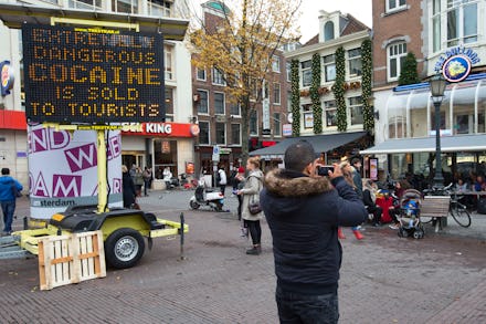 A tourist taking photos in Amsterdam with a sign reading 'Extremely dangerous cocaine is sold to tou...