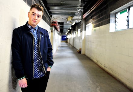 Sam Smith in a black jacket and blue shirt leaning on a wall in an industrial hall