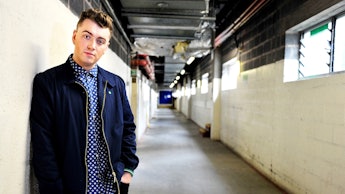 Sam Smith in a black jacket and blue shirt leaning on a wall in an industrial hall