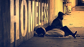 A homeless person with their sleeping bag sitting in front of the sign on the wall that says "HOMELE...