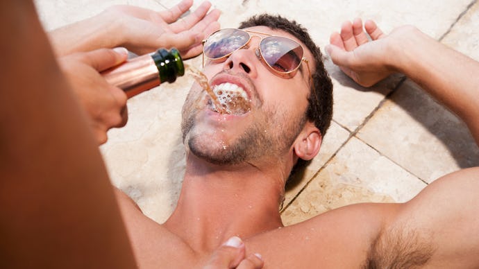 A person pouring champagne into a mouth of a man that is lying on the ground