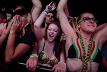 A crowd at an electronic music concert, with three girls at the front, two shouting and dancing and ...