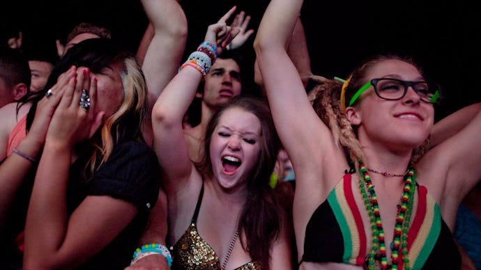 A crowd at an electronic music concert, with three girls at the front, two shouting and dancing and ...