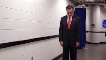 Mitt Romney standing in a black suite and red tie looking at the floor