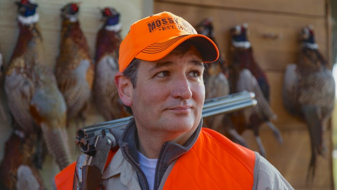 Ted Cruz with a hunting riffle on his shoulder and ducks hanging on a wall behind him