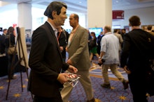Man wearing a Ronald Reagan mask in a black suite walking during a conference, holding promotional m...