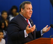 Chris Christie in a navy suit, a white shirt, and a red-blue tie with his arms up giving a speech in...