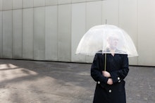 A trans person standing in a black coat and holding a see-through umbrella