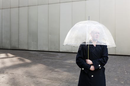 A trans person standing in a black coat and holding a see-through umbrella