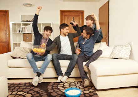 four men sitting on a couch with snacks cheering and fighting over the remote