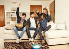 four men sitting on a couch with snacks cheering and fighting over the remote