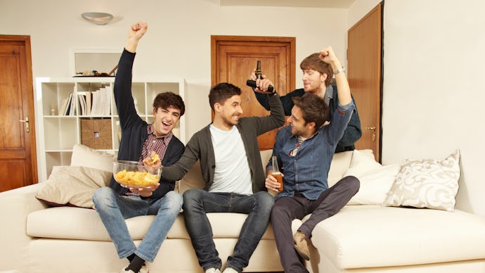 four men sitting on a couch with snacks cheering and fighting over the remote