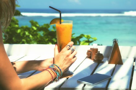 A woman sitting at the beach with her phone in front of her while holding a orange drink 