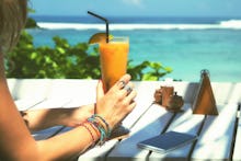 A woman sitting at the beach with her phone in front of her while holding a orange drink 