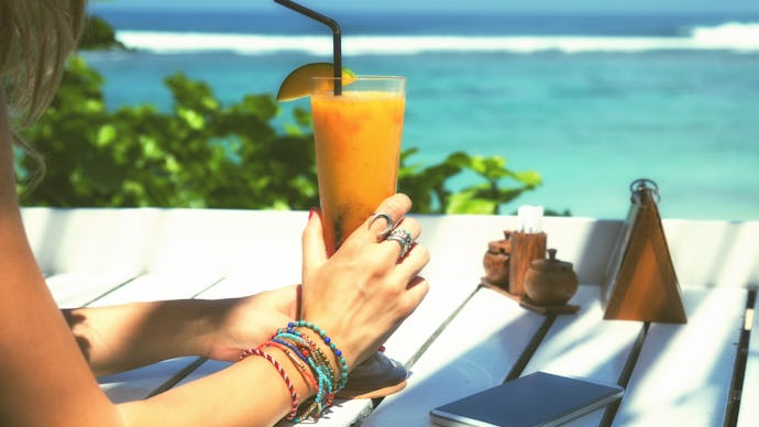 A woman sitting at the beach with her phone in front of her while holding a orange drink 