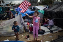 A girl waving an american flag in an immigration camp while other children run around 