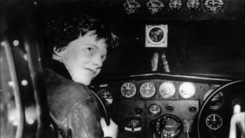 An old black and white image of Amelia Earhart sitting at the cockpit of a plane
