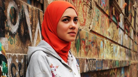 Mayam Mahmoud, an Egyptian rapper wearing a red hijab standing in front of a graffiti-covered wall