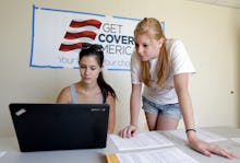 Girl with black hair sitting in front of the laptop, while a blond girl is standing next to her