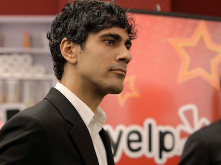 A man with curly dark hair in a black blazer, and a white shirt with a Yelp poster behind him