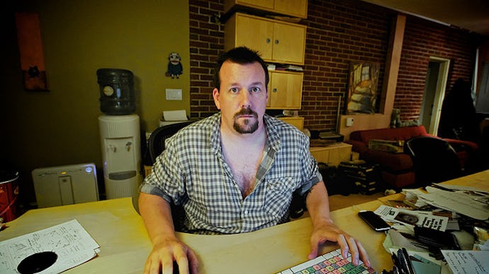 A man in a checkered shirt sitting at his work desk, with his e-mail on his computer, looking toward...