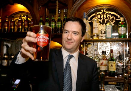 Man with short hair in black suite and tie standing in a pub holding a glass full of beer.