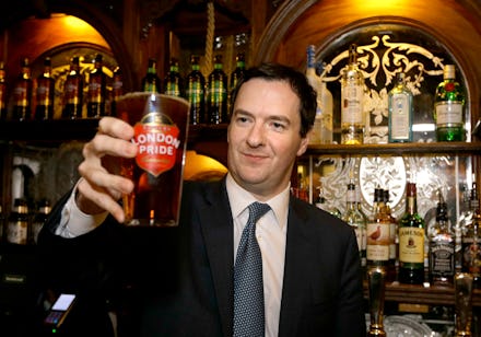 Man with short hair in black suite and tie standing in a pub holding a glass full of beer.