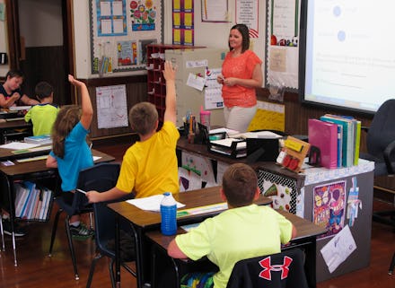A teacher in an orange top is in her classrom, students are raising their hands to answer a question