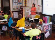 A teacher in an orange top is in her classrom, students are raising their hands to answer a question