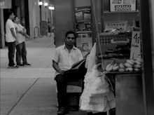 Black and white photo of an entrepreneur next to his food truck in the street, sitting on a milk cra...