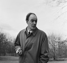 Black and white photo of Roald Dahl in a trench coat, smoking a cigarette