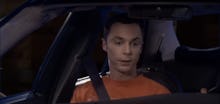 Sheldon Cooper in an orange shirt sitting in the passenger seat of a car