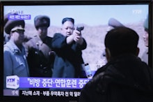 A man watching Kim Jong-un on tv, where he is holding a gun, and his companions standing on his side...