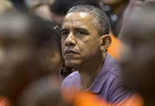 Former President Barack Obama sitting in a crowd wearing a blue button up shirt looking worried