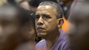 Former President Barack Obama sitting in a crowd wearing a blue button up shirt looking worried