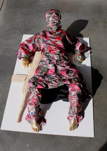 A person with gold hands and feet, in a pink and gray body suit, with a knitted gun, laying on a whi...