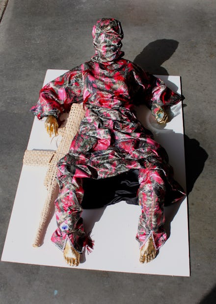 A person with gold hands and feet, in a pink and gray body suit, with a knitted gun, laying on a whi...