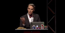 Bill Nye standing in front of a podium with a laptop in front of him giving a speech at a creationis...