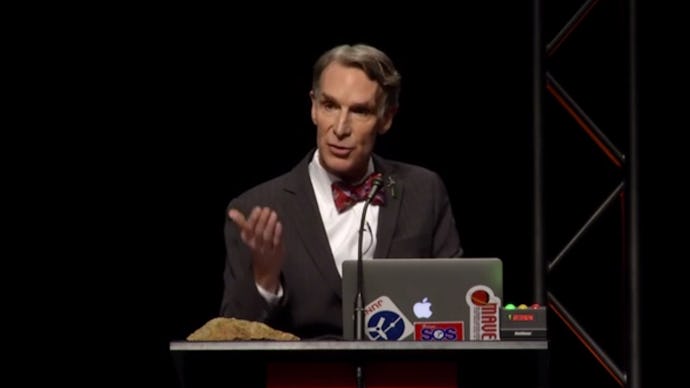 Bill Nye standing in front of a podium with a laptop in front of him giving a speech at a creationis...