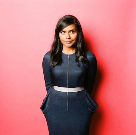 Mindy Kaling posing against a red wall, in a dark blue dress with a zip down the middle and a white ...