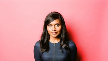 Mindy Kaling posing against a red wall, in a dark blue dress with a zip down the middle and a white ...