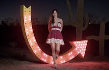 Kacey Musgraves in a red, flowy dress, standing with her hands crossed behind her back and a lit up ...