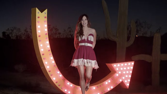 Kacey Musgraves in a red, flowy dress, standing with her hands crossed behind her back and a lit up ...